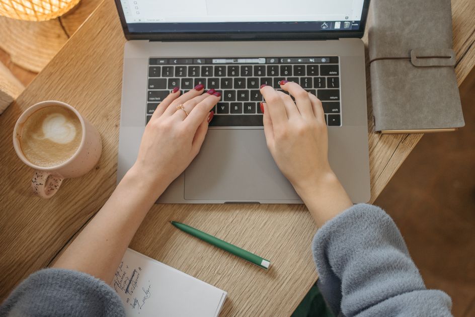 A person typing on a laptop with a notebook and coffee nearby