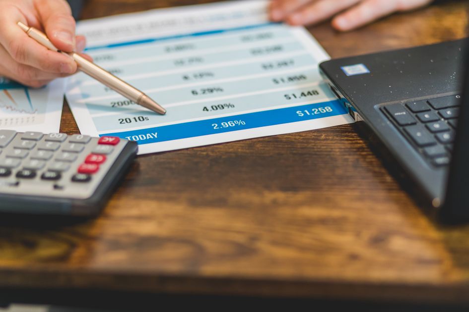 A hand pointing rates with a pen near a calculator and laptop