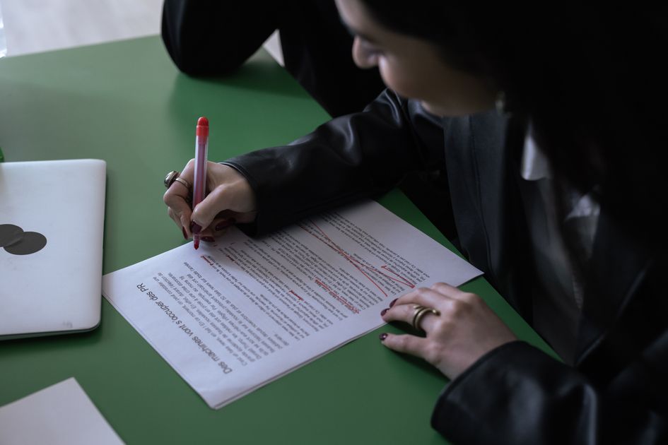 A woman using a red pen to edit a printed document beside a laptop