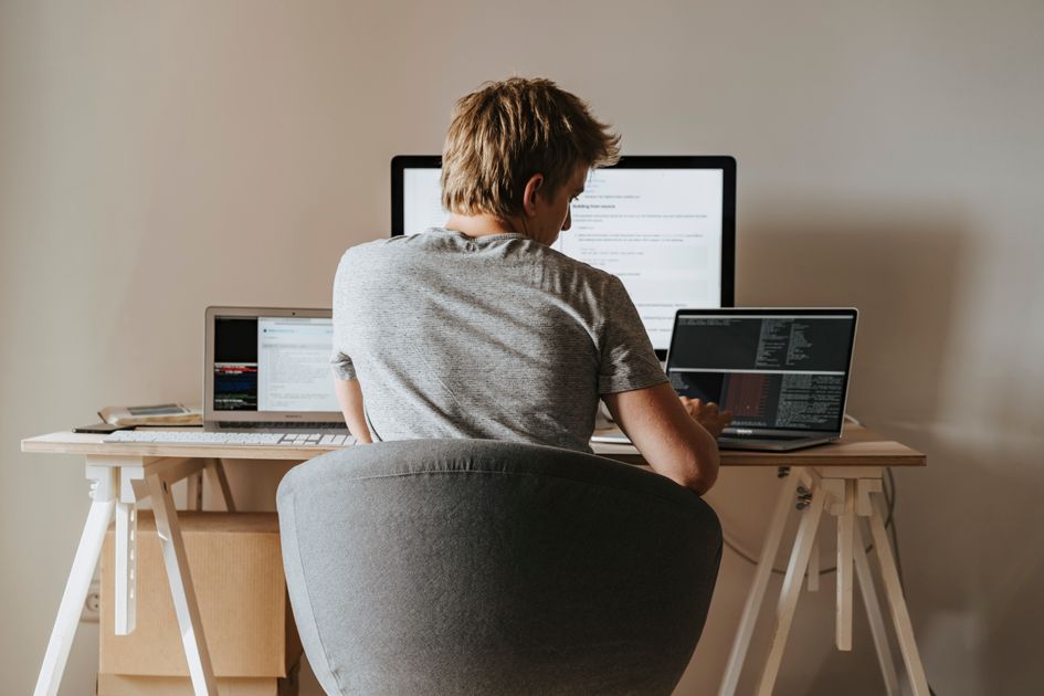 A young man coding at a home office with multiple laptops