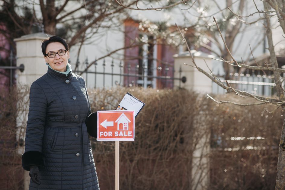 A smiling real estate agent standing with documents by a sign