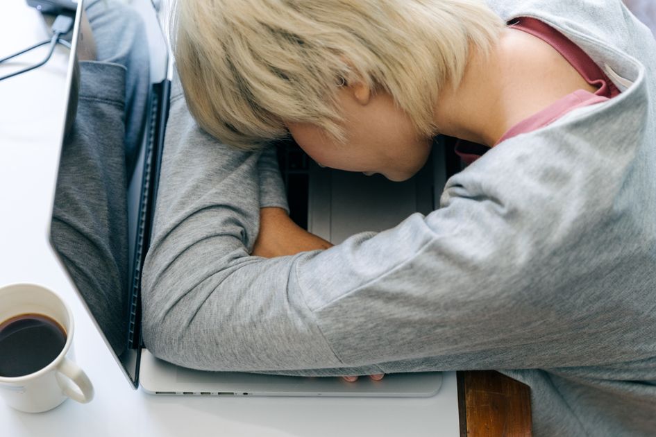 A woman resting her head on a laptop keyboard near a cup of coffee