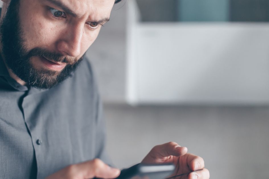 A bearded man looks shocked while holding a smartphone indoors