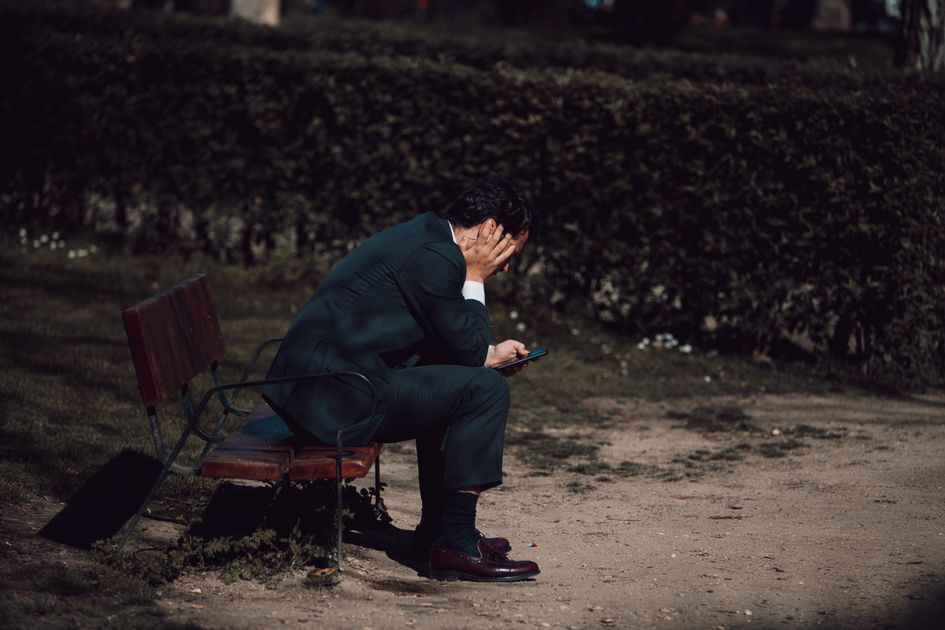 A worried man sits on a park bench holding a smartphone