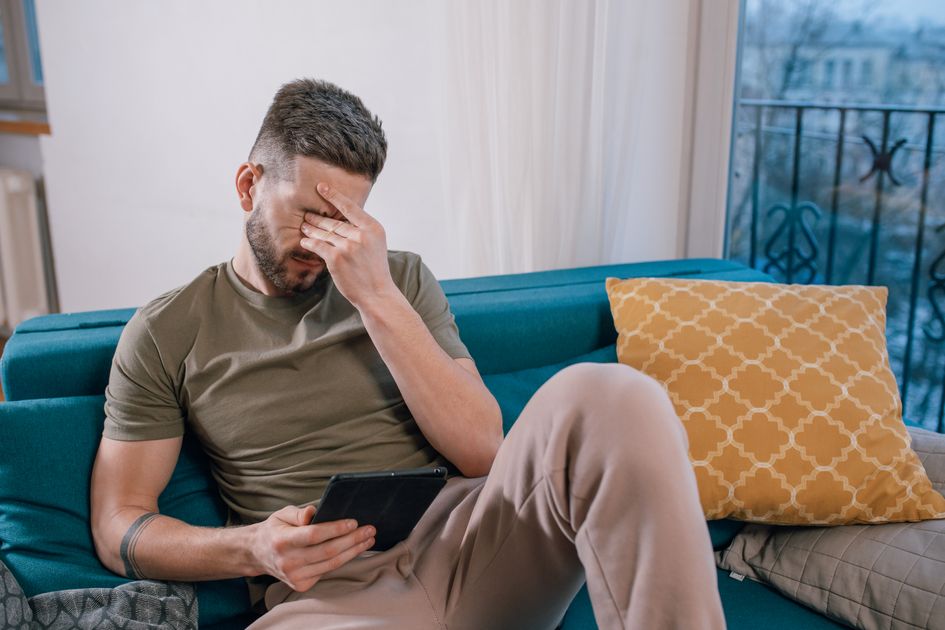 A man sits on a sofa holding a tablet, visibly stressed