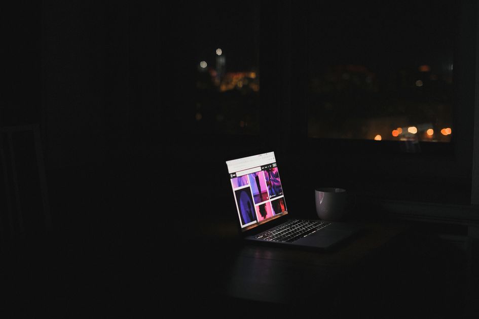 A laptop glows in a dark room at night with a cityscape through the window