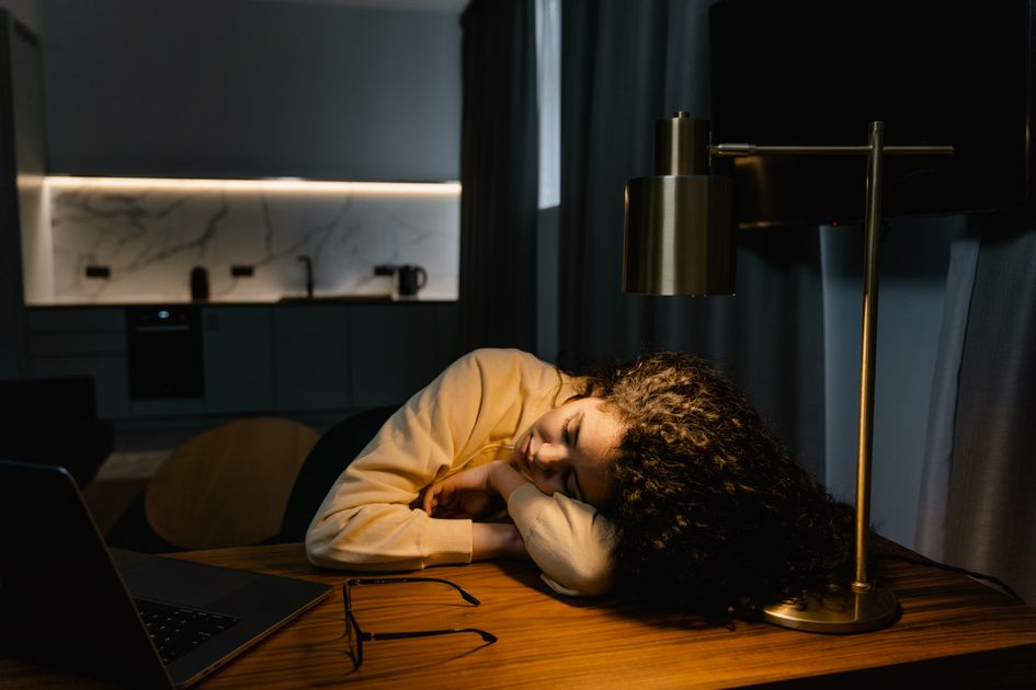 A tired person asleep at their desk under lamp light