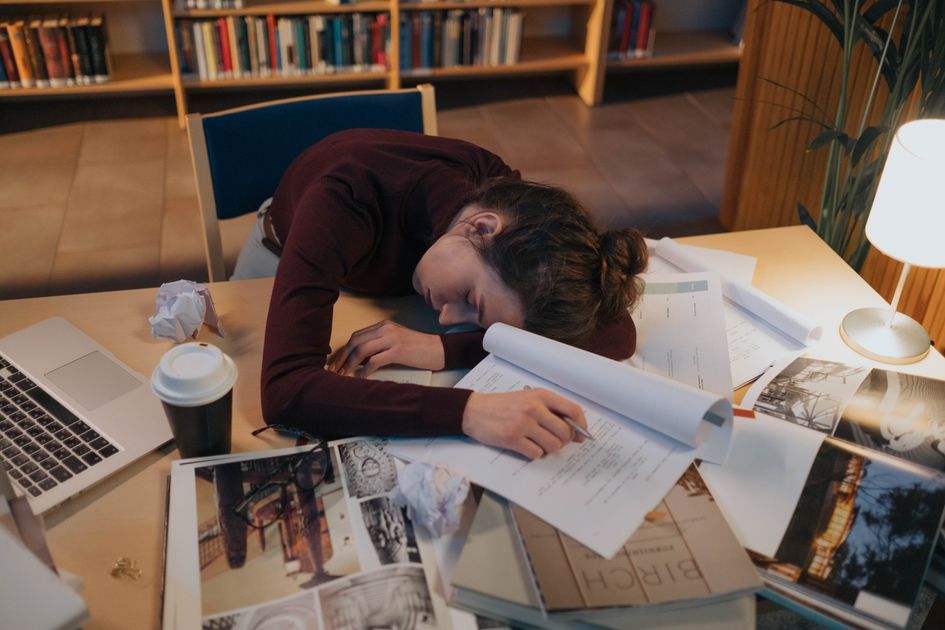 A tired person asleep at a desk covered in papers